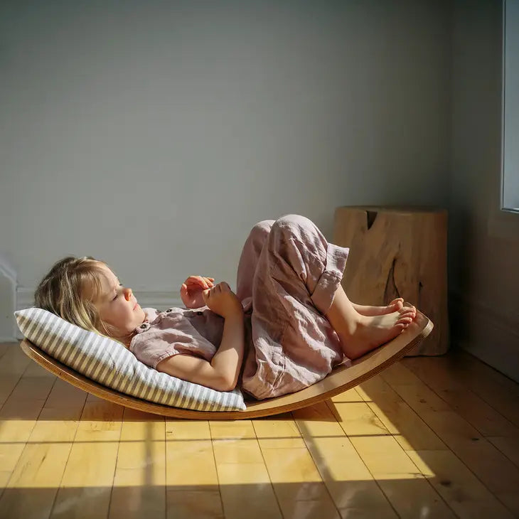 A child with light skin tone and blonde hair lays on the Kinderboard Balance Board. There is a pillow under their head and their knees are bent, with their feet resting on the end of the board.