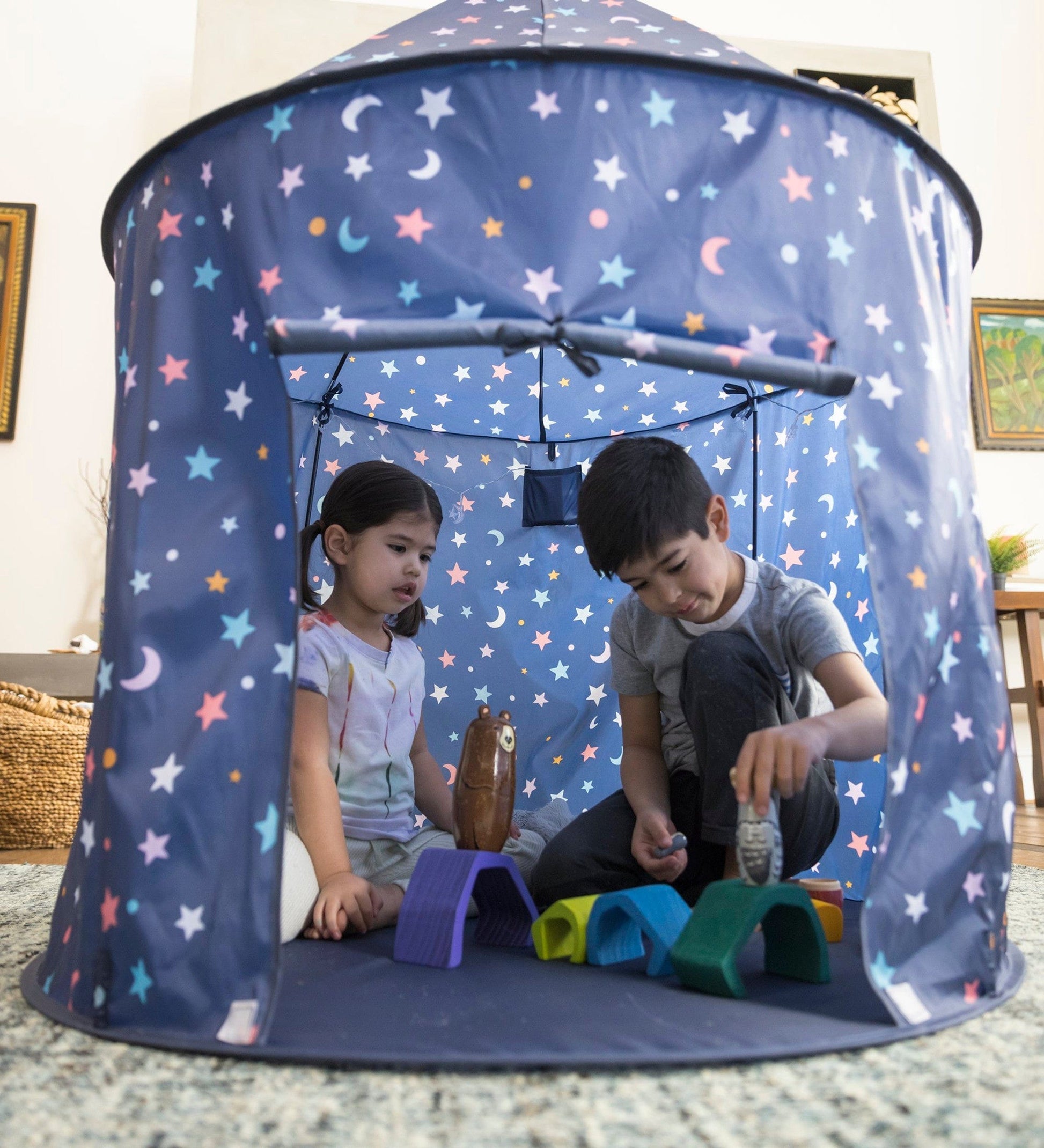 Two children sit inside the Light-Up Celestial Play Tent and play with several toys.