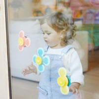 A child with light skin tone and short. curly dark blonde hair stands in front of a sliding glass door. The three varieties of Sensory Spinners are stuck to the glass, and the child is looking closely at one of them with one hand outstretched. 