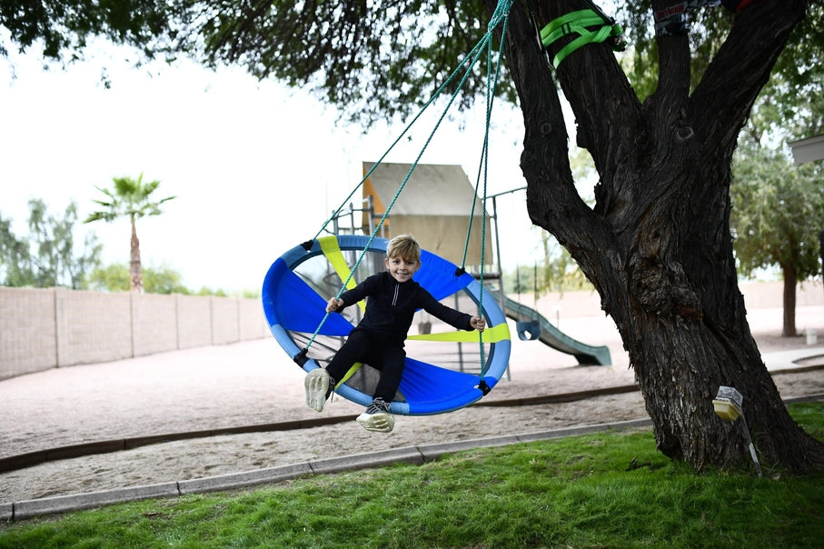 A child with light skin tone and short blonde hair swings back with their legs out in front of them on the Slackers 50" Adventure Sky Swing.