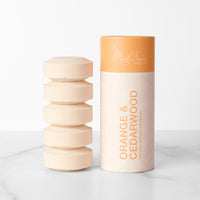 A stack of five orange Shower Steamers sit next to the product package for the Orange & Cedarwood variant.