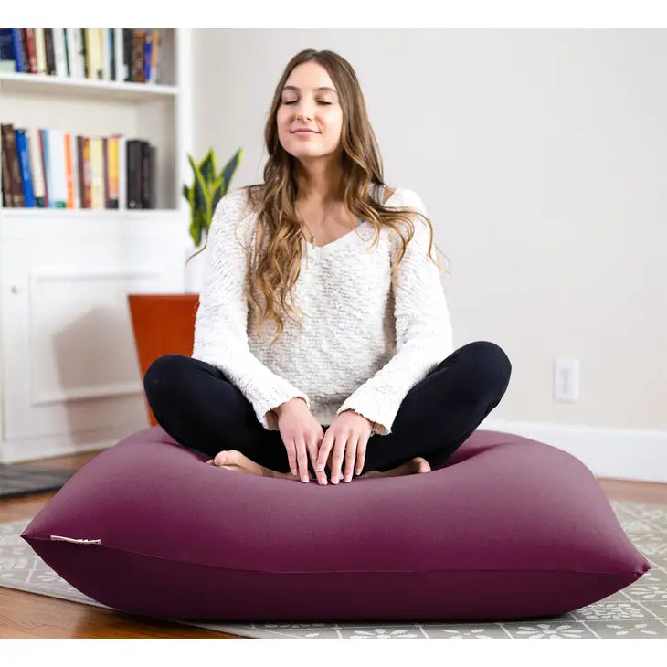A person with long brown hair sits with their legs crossed on top of the Deep Purple Yogibo Mini.