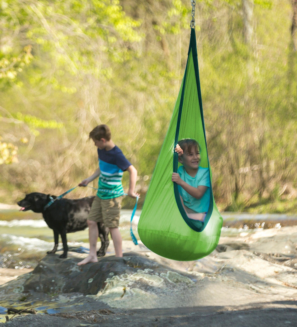 A child is hanging in the green HugglePod Lite Nylon Hanging Chair in a river spot. A child in the background is holding onto the leash of a black lab.