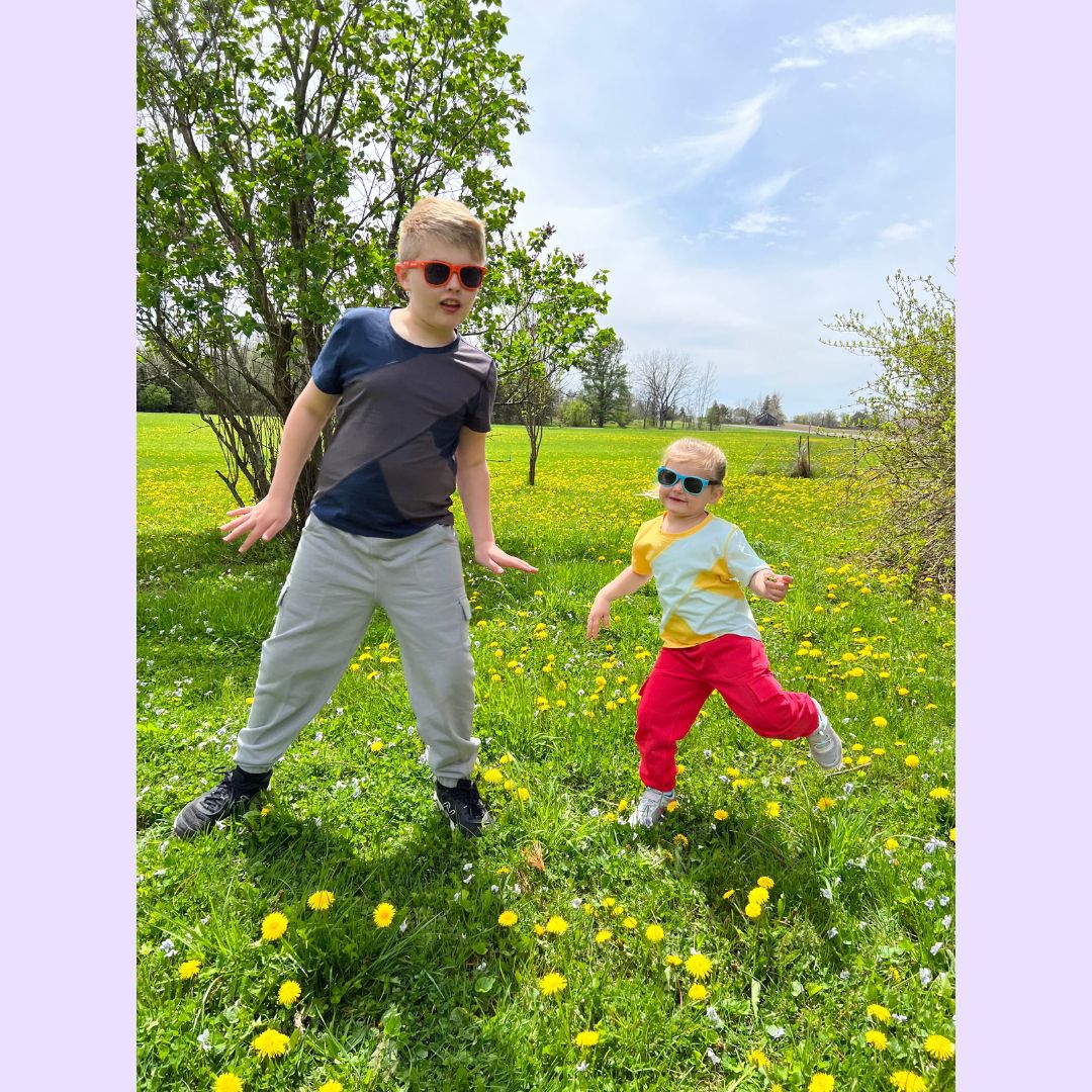 One small and one tall child stand in a green field full of yellow dandelions in the two versions of the Easy to See Sensory Friendly T-Shirt.