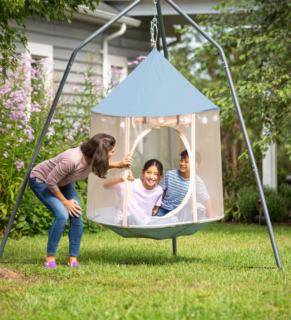 An adult looks into the HugglePod Panorama HangOut, where two children are sitting. They are in the yard.