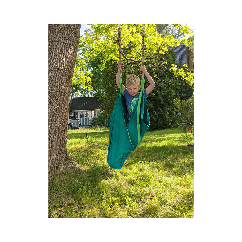 A child with light skin tone and short blonde hair is standing in the Slackers Swing Hammock.