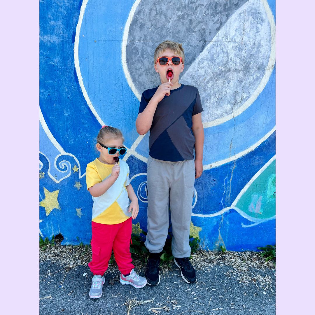One small and one tall child wear sunglasses and eat lollipops while wearing both versions of the Easy to See Sensory Friendly T-Shirt.