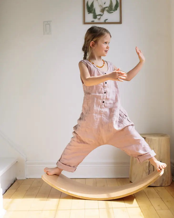 A child with light skin tone and a light brown braid stands on the Kinderboard Balance Board. Either leg is extended to opposite sides of the board, and their arms are held up as though balancing.