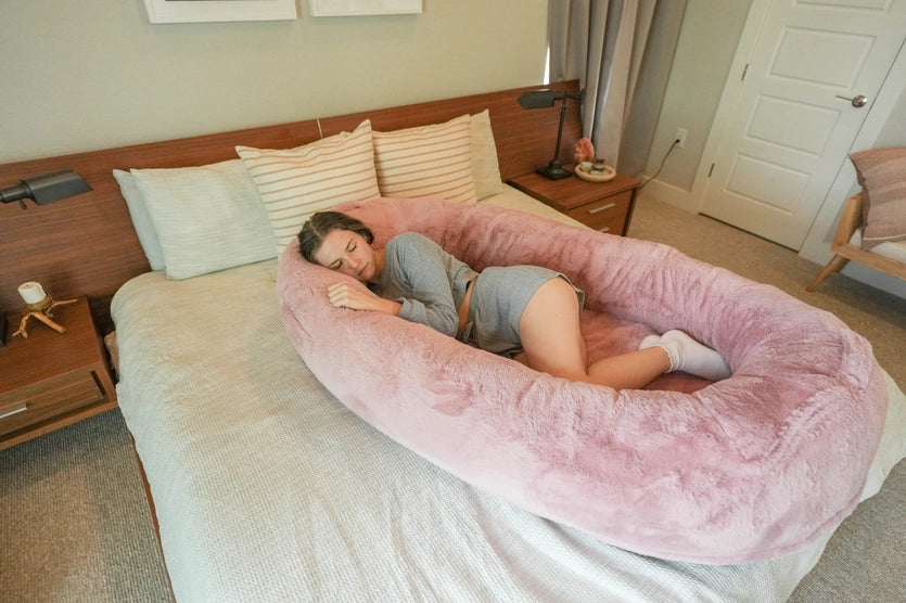A person lays in a pink Human Dog Bed on top of a regular bed in a bedroom.