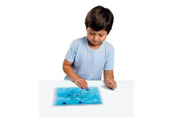 A child with light skin tone and short dark brown hair sits in front of the Tic-Tac-Toe Gel Pad. They are pushing one of the blue marbles.