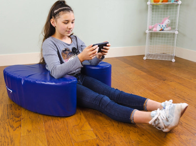 A child with light skin tone and a long brown ponytail sits on the wooden floor, positioned inside of the Sensory Soft Squeeze Seat by Bouncyband. They are playing a handheld videogame.