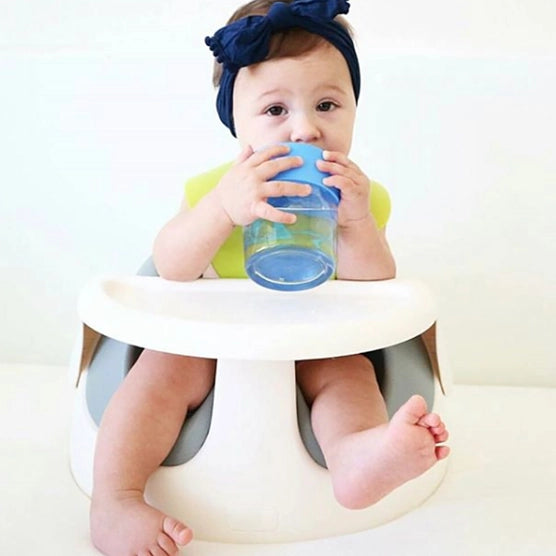 A toddler with light skin tone and short brown hair is sitting in a babyseat and holding the blue Stretchy Silicone Lid up to their face to take a drink from a clear cup.