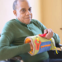 An older person with dark skin tone and very short grey hair relaxes in a wheelchair. Their left hand is inside of the TwiddleNathan and the other is moving a fidget attached to the outside.