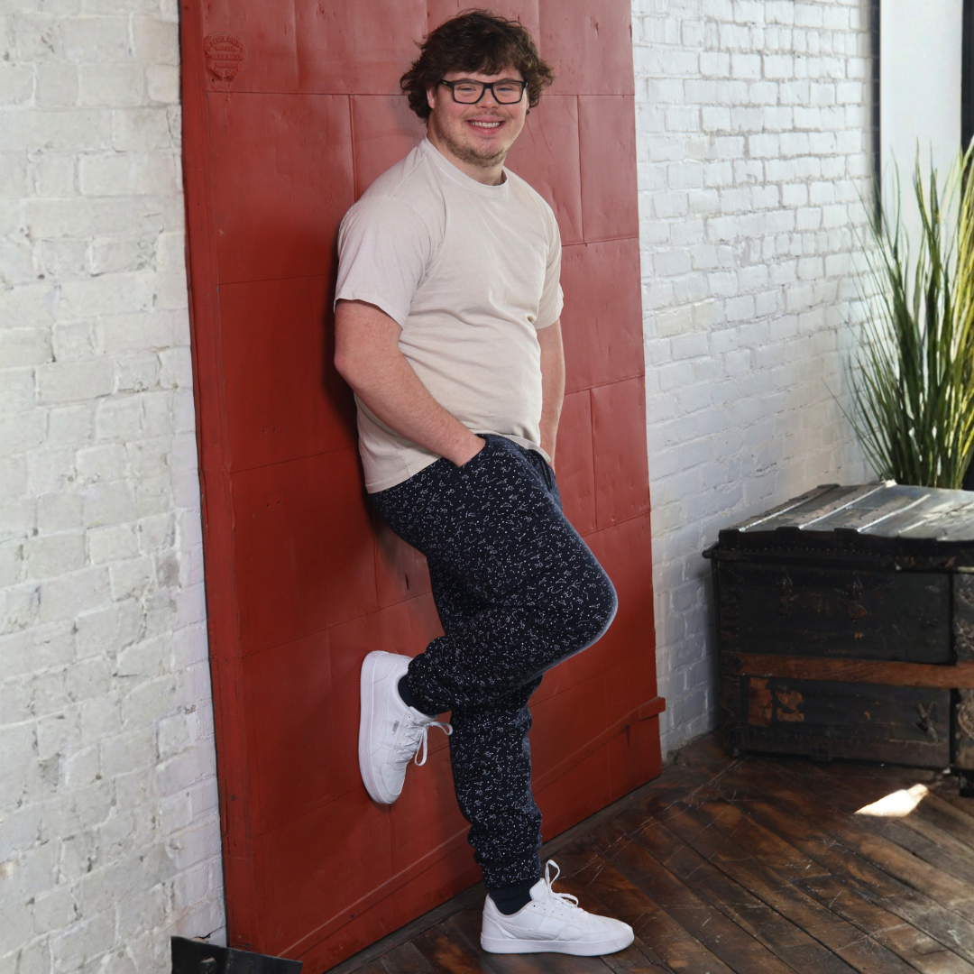 An adult smiles and leans back on a sliding barn door to model the Constellation Sensory Friendly Fidget Joggers.