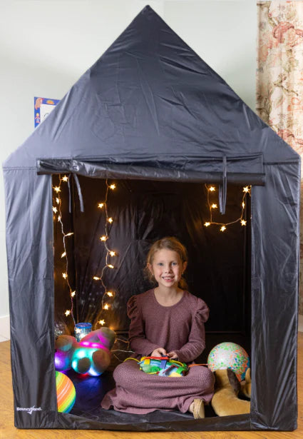 A child with light skin tone and a dark blonde ponytail sits with their legs crossed inside the Indoor Framed Dark Den. There are white lights on a string behind them and several glowing plushes on the ground around them.