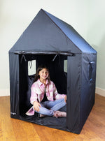 A child with light skin tone and long dark brown hair sits inside the Indoor Framed Dark Den with their legs crossed, smiling.