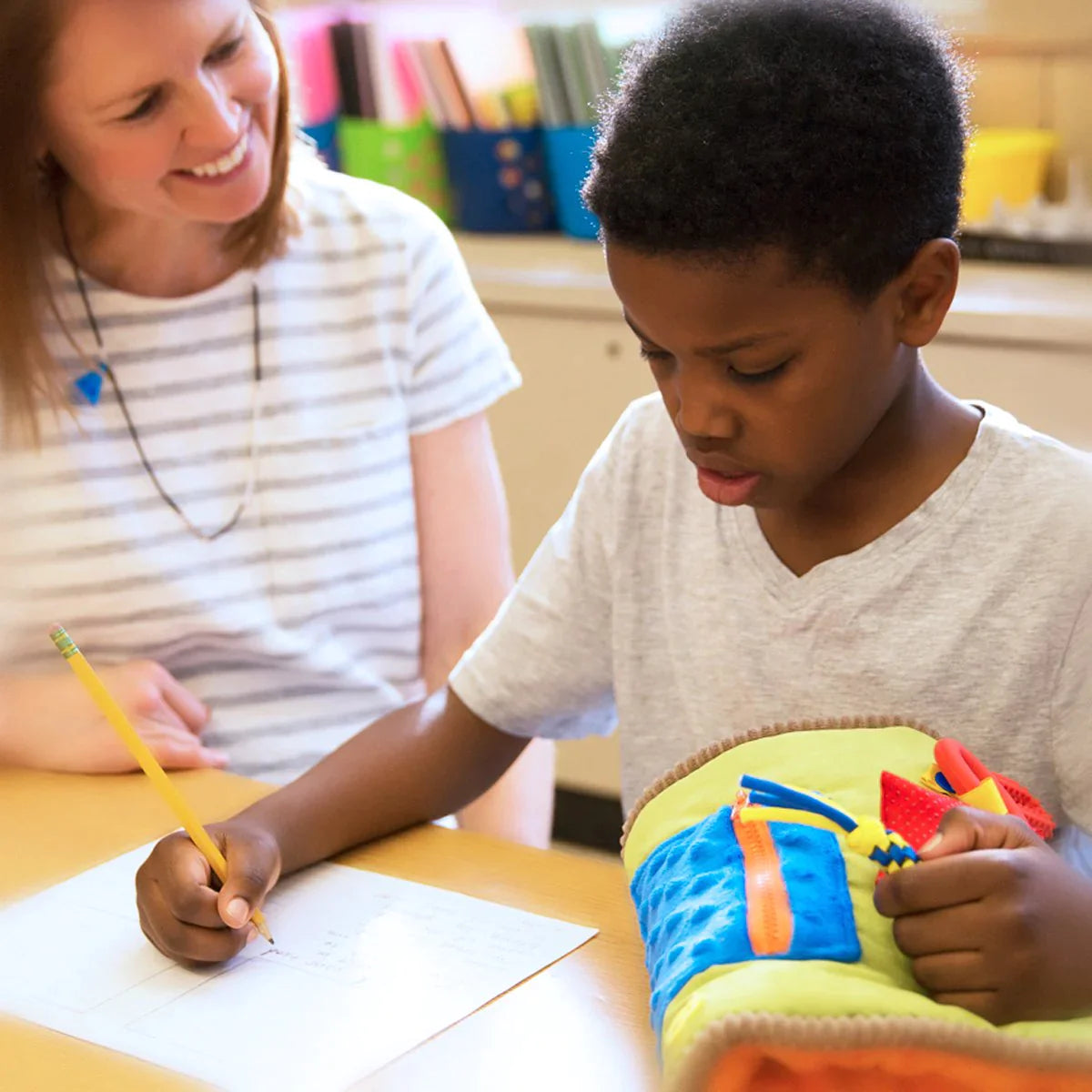 A child with dark skin tone and short black hair is sitting at a desk. They are writing with one hand and holding onto a fidget on the TwiddleNathan with the other. An adult sits next to the child smiling at him.