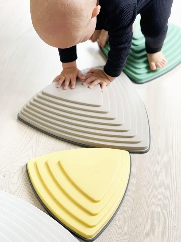 A toddler with light skin tone stands on a Gonge River Stones Nordic and is bent over, placing their hands on another stone.