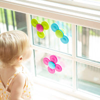 A child with light skin tone and short blonde hair stands in front of a sunlit window while looking at the colorful Whirly Squigz.