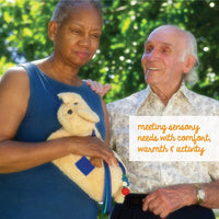Someone with dark skin tone and short grey hair is holding onto the soft TwiddlePup. A man with light skin tone and short grey hair is standing next to them and smiling.