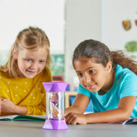Two children sit at a desk with two books open in front of them on the table. They are both watching the sand fall in the Jumbo 10 Minute Sand Timer.