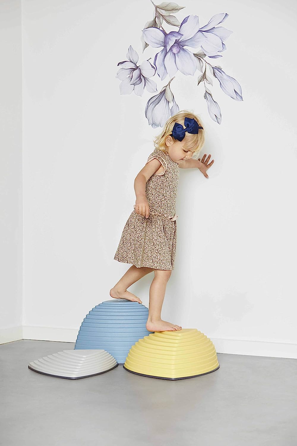 A child with light skin tone and short blonde hair with a blue bow steadiness themself on the side of the wall and moves from the tallest Gonge Hilltop Nordic to the medium sized stone.