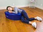 A child with light skin tone and a long brown ponytail lies on a wooden floor and onto the Sensory Soft Squeeze Seat by Bouncyband.