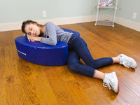 A child with light skin tone and a long brown ponytail lies on a wooden floor and onto the Sensory Soft Squeeze Seat by Bouncyband.