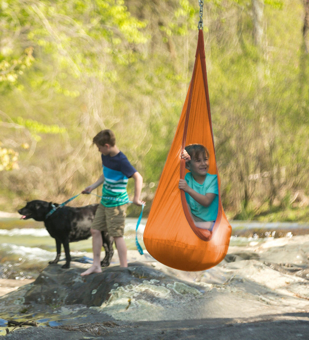 A child sits in an orange HugglePod Nylon Hanging Chair. A child in the background is holding onto the leash of a black lab.