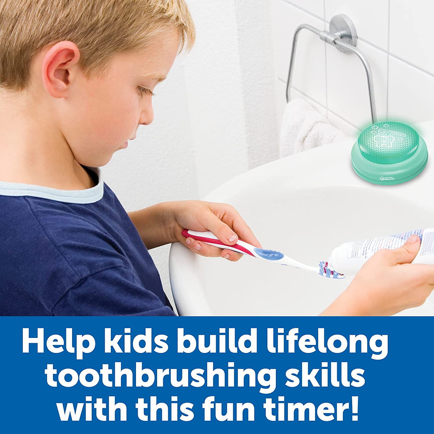 A child with light skin tone and short blonde hair puts toothpaste on a toothbrush over a bathroom sink. There is a Toothbrush Timer on the sink ledge.