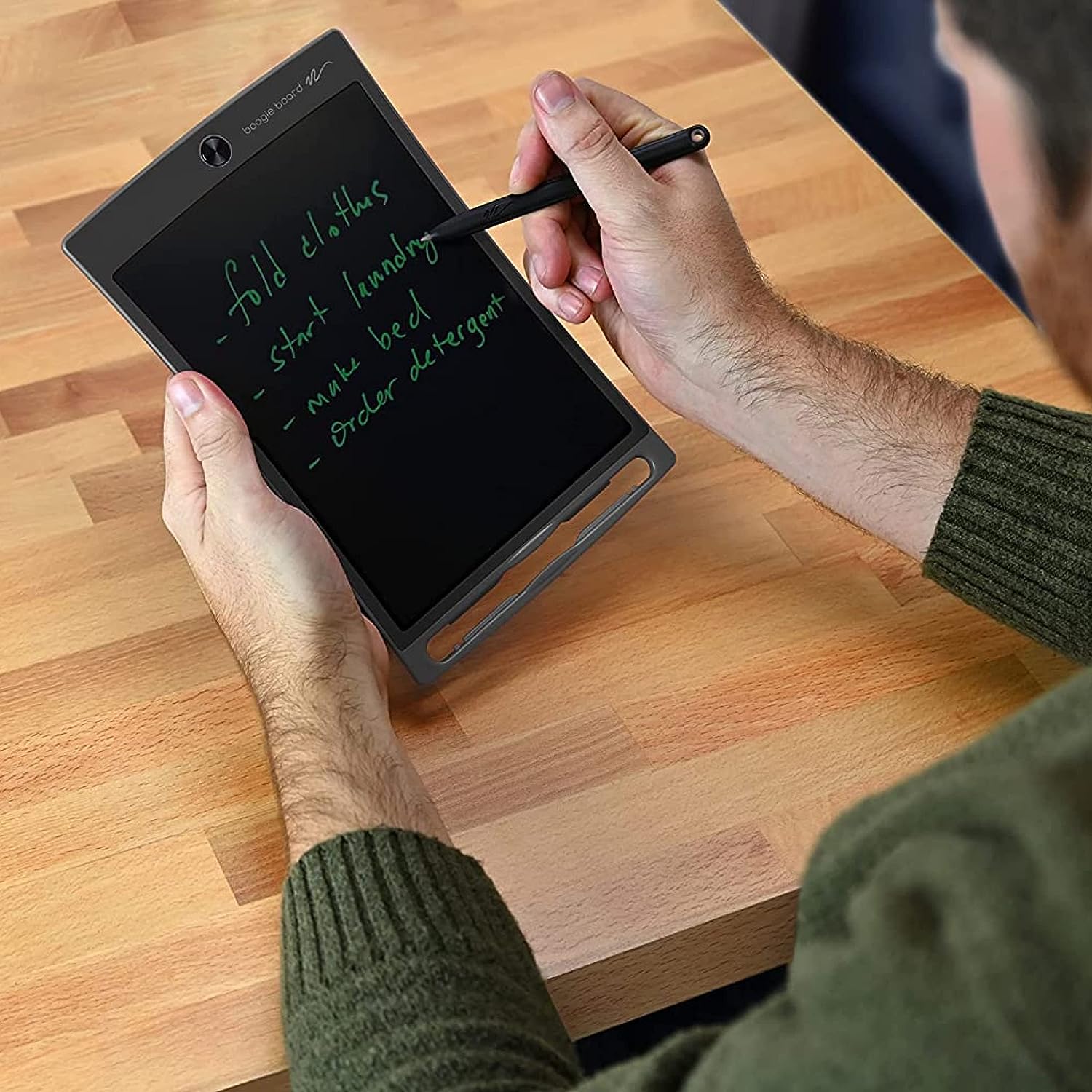 A person with light skin tone sits at a wooden table holding onto the Boogie Board Jot.