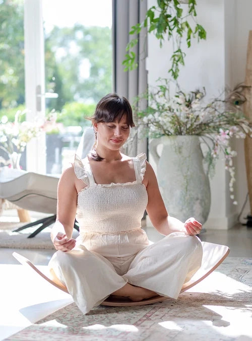 An adult with light skin tone and brown hair pulled back in a braid is sitting cross legged in the middle of a Kinderboard. Their hands are in meditative positions above their knees and their eyes are closed.