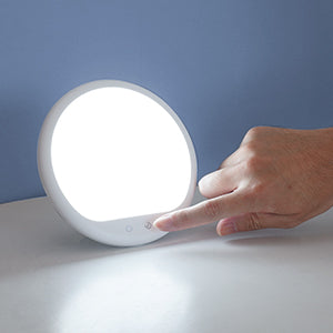 A hand with light skin tone reaches for the brightness button on the Light Therapy Lamp.