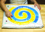 An adult with light skin tone and pink nail polish is pointing at a marble in the Spiral Gel Pad.