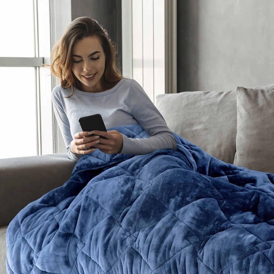 A person with light skin tone and long brown hair leans back on a couch while looking at their phone. They are covered in the Dream Theory Butter Velvet Weighted Blanket 15 lb.