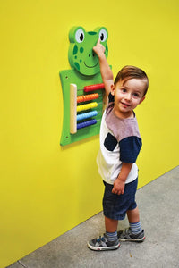 A child with light skin tone and short brown hair points to the eye of the frog in the Frog Activity Wall Panel.