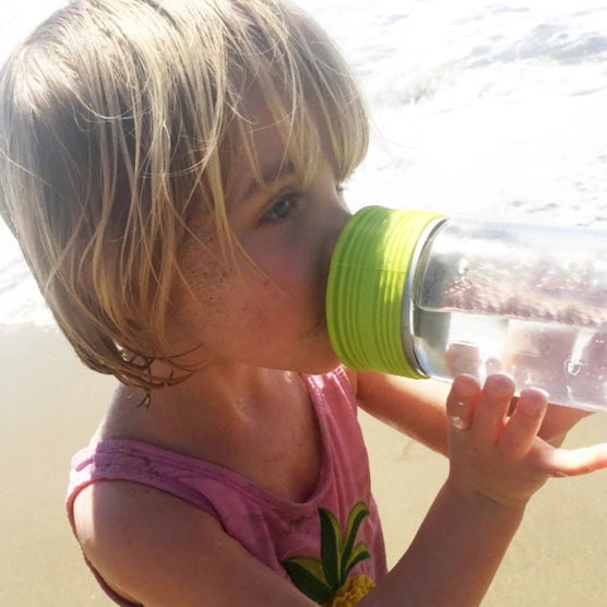 A child with light skin tone and shoulder-length blonde hair is holding up a cup with the lime green Stretchy Silicone Lid, and taking a drink from it.