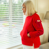 An older person with light skin tone and shoulder length blonde hair puts their hands in the pockets and looks out of an open window contentedly.