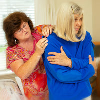 An older person with light skin tone and shoulder length blonde hair crosses their arms across their chest while their caretaker buttons a blue AdaptiWrap at the shoulder.