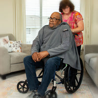 An older person with dark skin tone and receding hair is sitting in a wheelchair while a caretaker secures a grey Adaptiwrap around their shoulders.