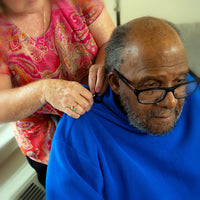 An older person with dark skin tone who is balding is having the blue AdaptiWrap buttoned on the shoulder by a caretaker.