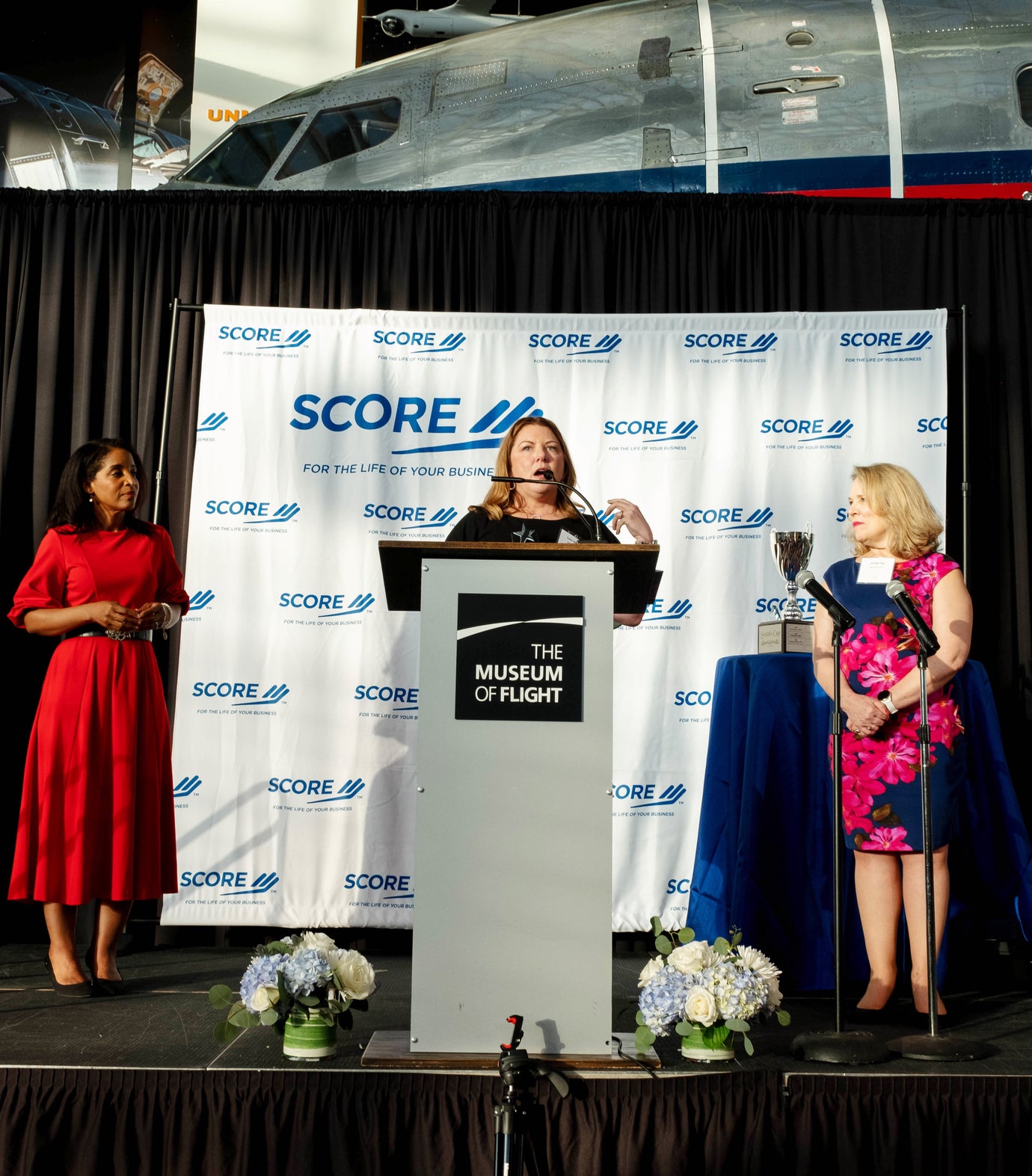 Angela Poe Russell, Katie McMurray, and Jennifer Dye standing on stage while Katie speaks to an audience at the podium