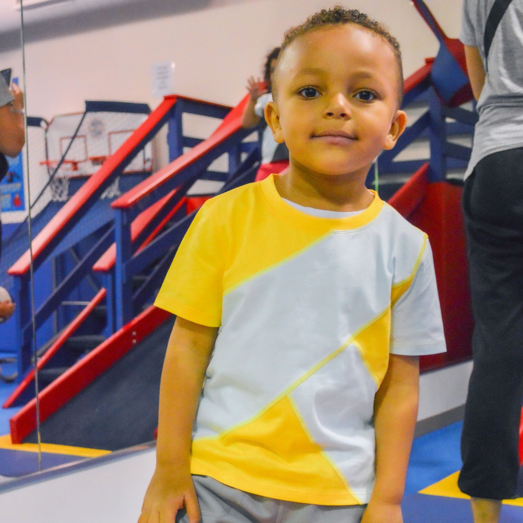 A child grins while wearing the Yellow and Mint Sensory Friendly T-Shirt at a gym.