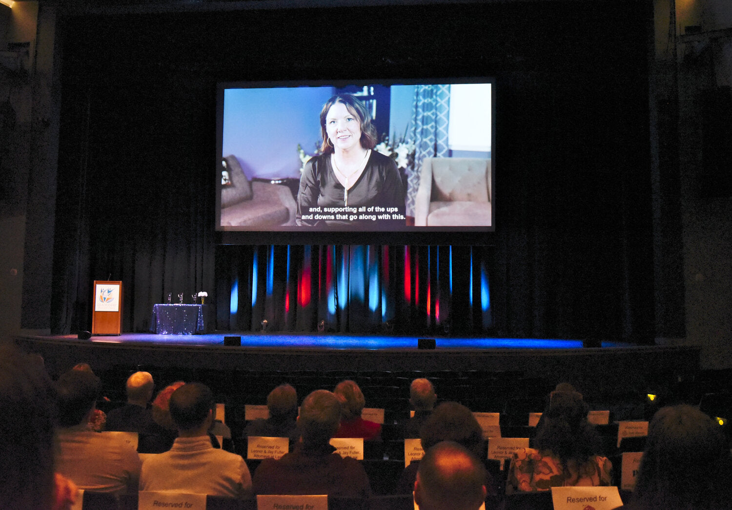 filled theater seats with people watching a video on stage of a woman talking. Closed captioning says "and supporting all of the ups and downs that go along with this"
