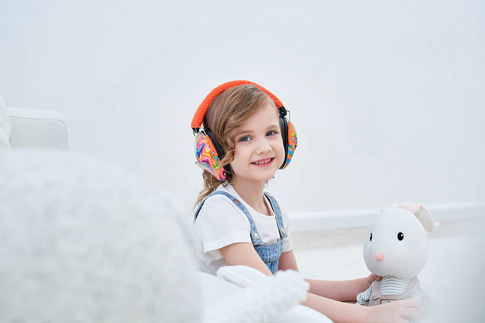 A child with light skin tone and shoulder-length curly brown hair is looking directly at the camera. They are holding a white stuffed animal and wearing the Lollipop Noise Reduction Headphones.