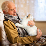 An elderly man covered in a blanket holds the Therapy Cat Plush on his chest.