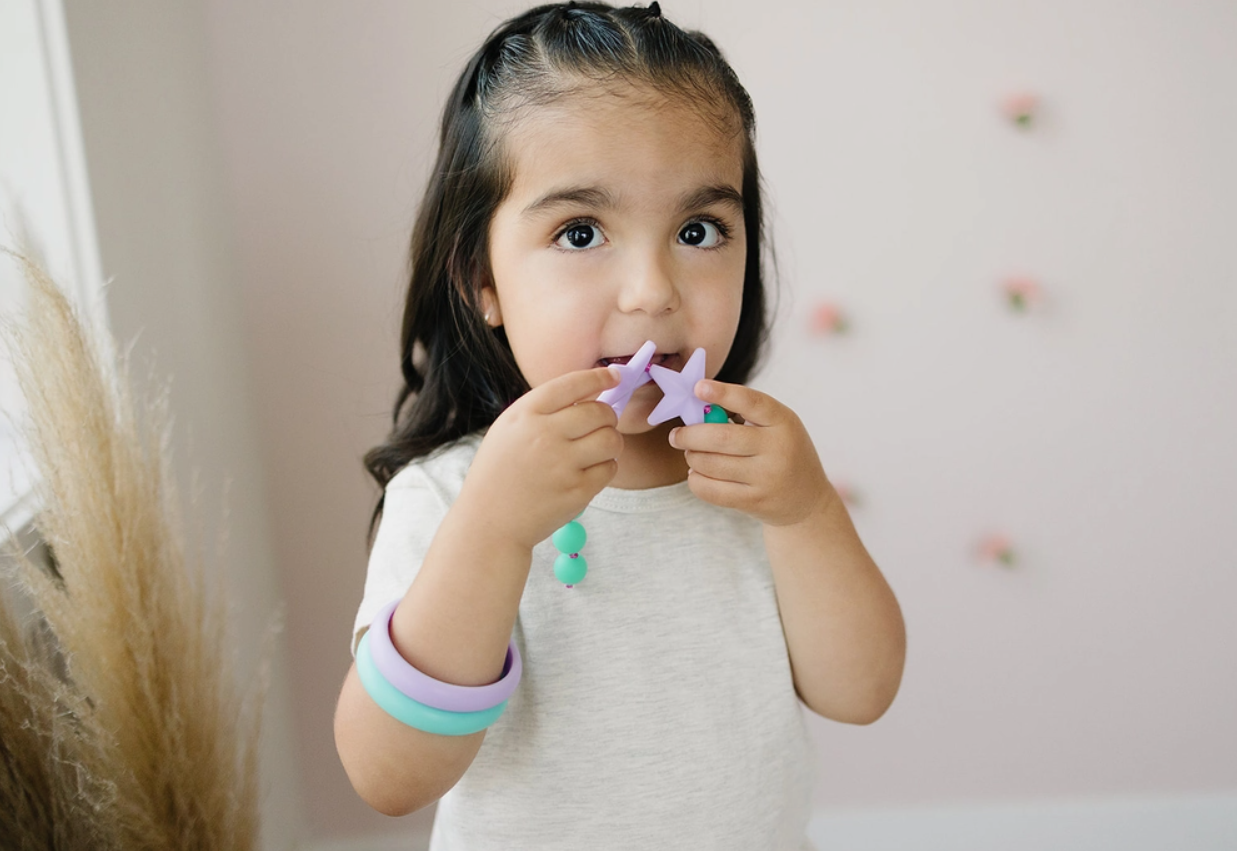 A child with medium skin tone and long dark hair is looking up and holding the lavendar Juniorbeads Broadway Star Necklace up to their mouth.