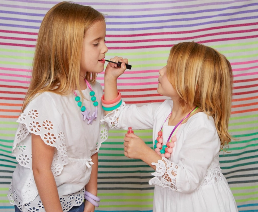 Two children are standing next to each other. One of them is wearing the lavendar Juniorbeads Broadway Star Necklace while the other child applies lipgloss to their lips.