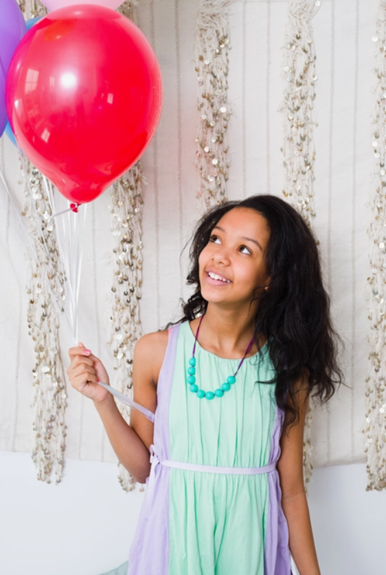 A child with dark skin tone and long dark hair is holding several balloons and wearing the spearmint Juniorbeads Madison Jr. Necklace.