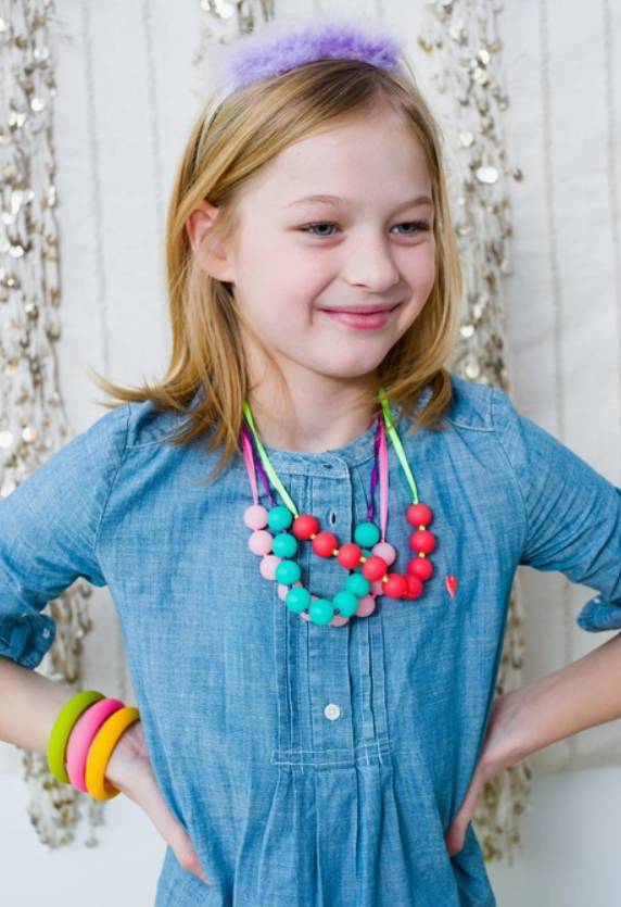 A child with light skin tone and shoulder length blonde hair is wearing several different colors of the Juniorbeads Madison Jr. Necklace. They are wearing a fuzzy purple tiara and their hands are on their hips.